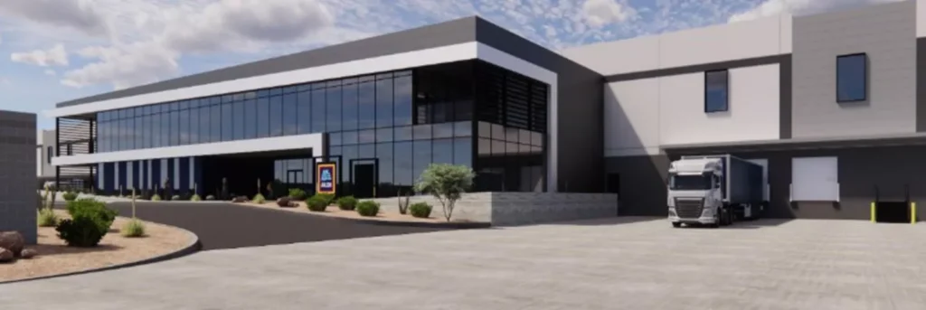 Architectural rendering of a large modern commercial facility with floor-to-ceiling glass curtain wall facade, dark and light grey cladding, desert landscaping, and a semi-truck at a loading dock on the right side; an Aldi logo sign is visible near the entrance.