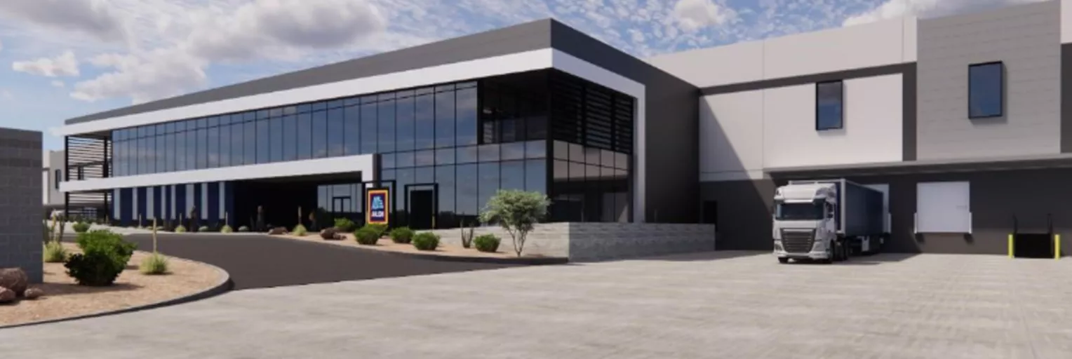 Architectural rendering of a large modern commercial facility with floor-to-ceiling glass curtain wall facade, dark and light grey cladding, desert landscaping, and a semi-truck at a loading dock on the right side; an Aldi logo sign is visible near the entrance.