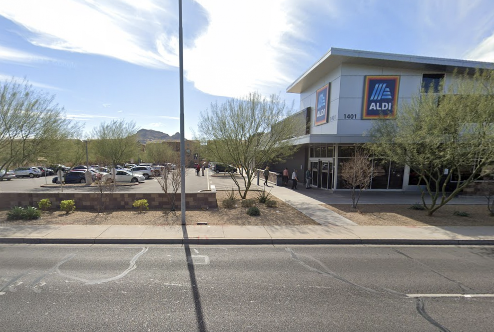 An ALDI grocery store with its blue-and-orange logo prominently displayed on a modern white facade, set in a desert-landscaped strip mall parking lot with palo verde trees and mountains visible in the background.