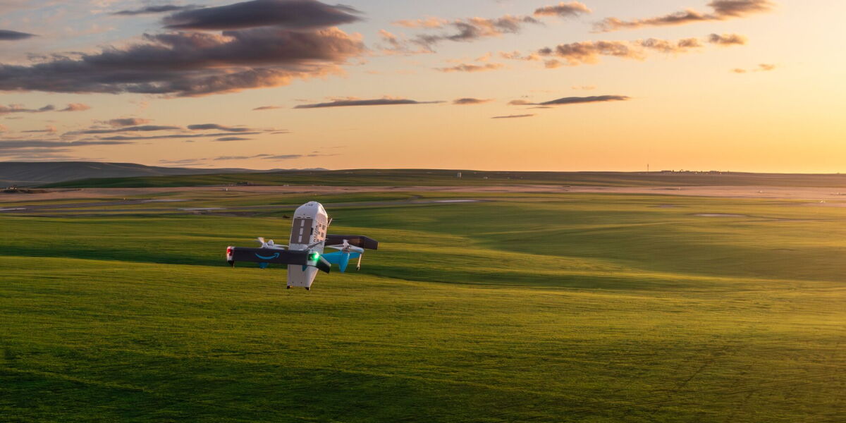 An Amazon Prime Air drone with the Amazon smile logo and teal-blue accents hovers low over a green field at golden-hour sunset.