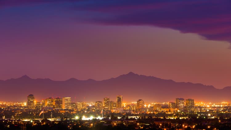 The Phoenix, Arizona skyline at dusk, showing a glowing urban core of mid-rise and high-rise buildings under a vivid purple-magenta gradient sky backed by a silhouetted mountain range.