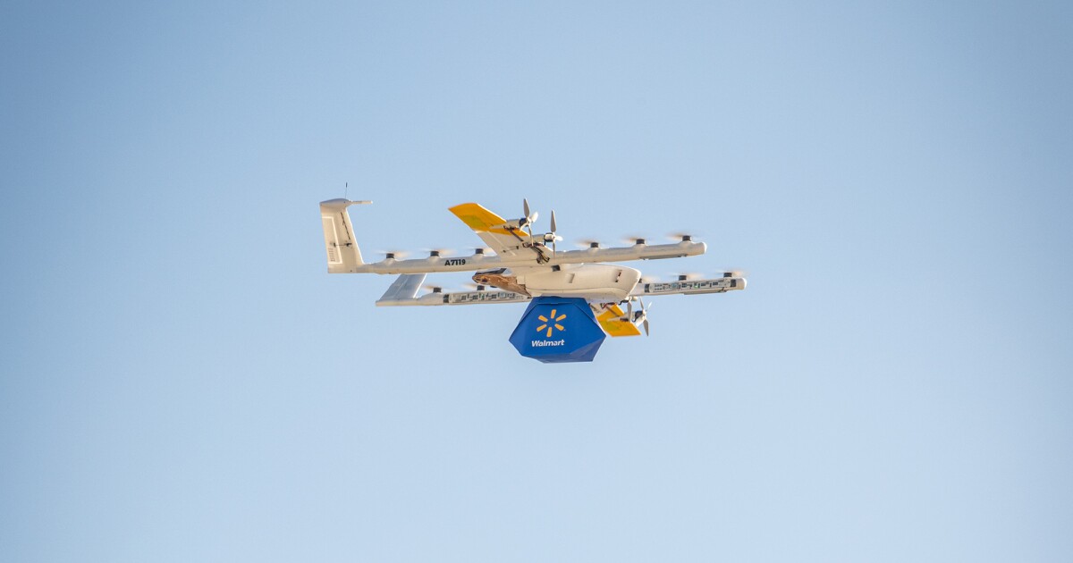 A white fixed-wing multirotor delivery drone labeled A7119 carrying a blue Walmart-branded package flies against a clear light-blue sky.