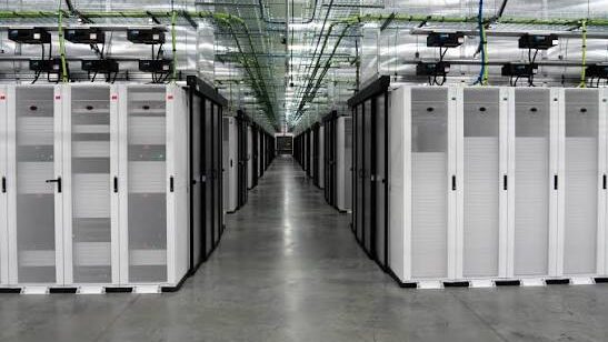 A long corridor flanked by rows of tall white server rack cabinets extending to a vanishing point inside a large data center, with overhead cooling and cable management infrastructure on the ceiling.