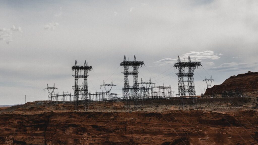 A large electrical substation with multiple high-voltage transmission towers and interconnecting power lines sits atop red sandstone cliffs under an overcast grey sky in an arid desert landscape.