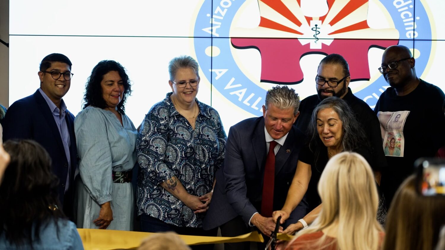Group of approximately seven people at a ribbon-cutting ceremony in front of a large screen displaying the Arizona Valley clinic logo, with two individuals in the foreground cutting a yellow ribbon while others smile and look on