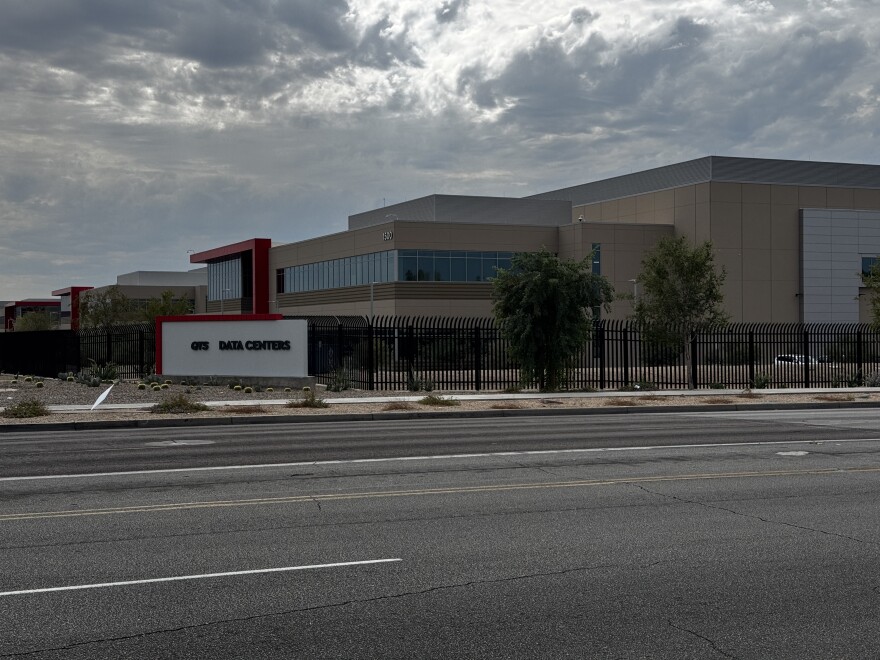 A large tan commercial data center building with red accent architecture behind a black iron security fence, viewed from across a multi-lane road in Arizona.