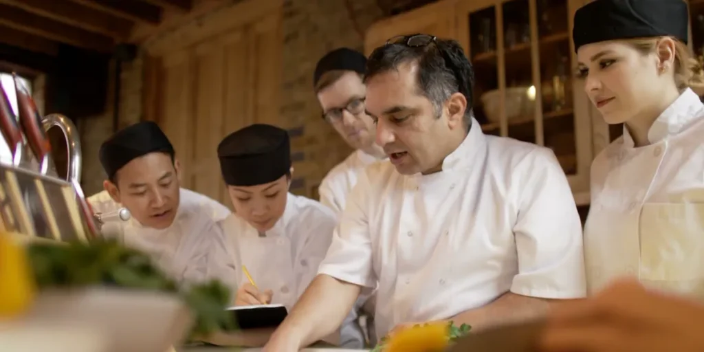 A head chef in a white double-breasted jacket with sunglasses on his head speaks to four culinary staff members in white jackets and black toques in a warm wood-paneled kitchen.