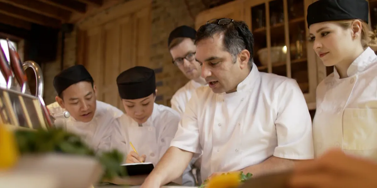 A head chef in a white double-breasted jacket with sunglasses on his head speaks to four culinary staff members in white jackets and black toques in a warm wood-paneled kitchen.