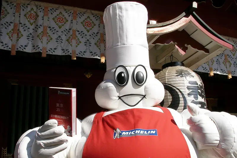 A large Michelin Bibendum mascot dressed as a chef with a white toque and red apron holds a red Michelin Guide book with Japanese text in front of a traditional Japanese shrine with paper lanterns.