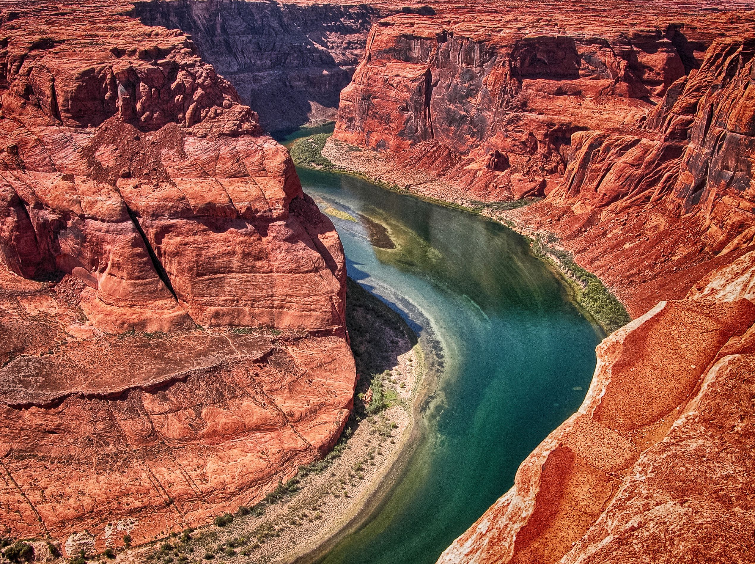 Aerial view of the Colorado River cutting through red sandstone canyon walls, with turquoise-green water curving through the gorge and sandy banks at the inner bend, near Page, Arizona
