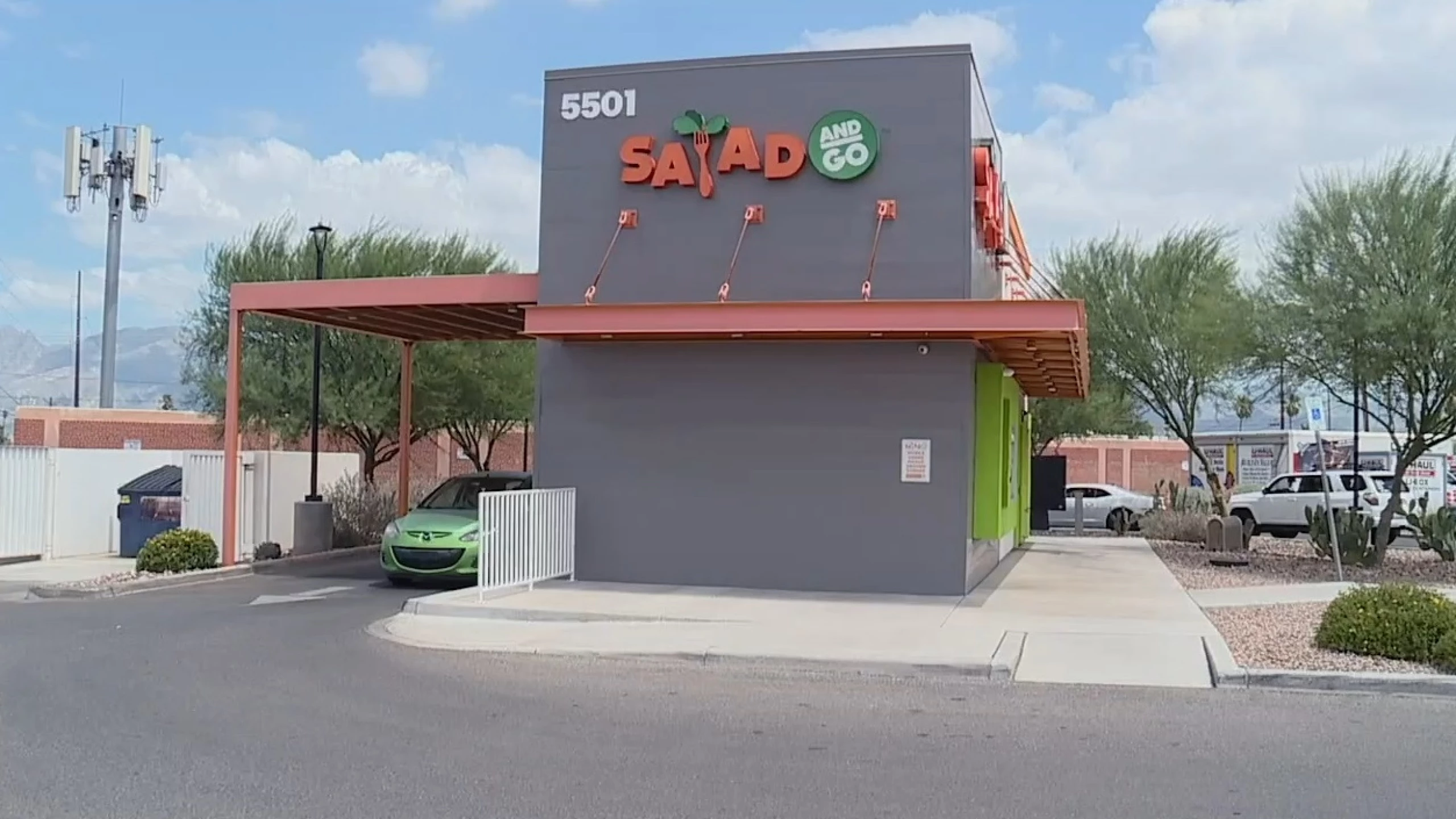 Exterior of a Salad and Go drive-thru restaurant showing brand signage, orange canopy, green accent panels, and a green Mazda in the drive-thru lane against a partly cloudy desert sky with mountains in the background