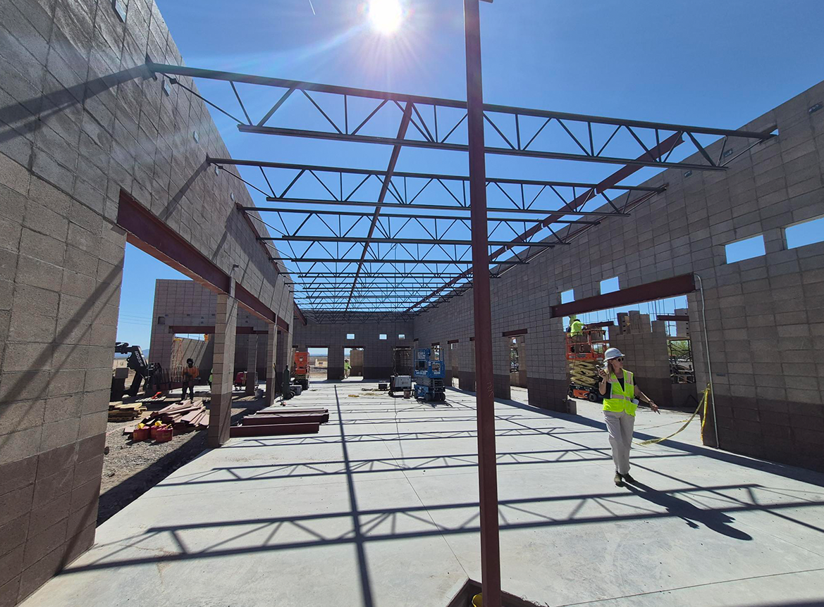 Interior of a large commercial building under construction showing concrete masonry walls, exposed steel roof trusses, a concrete slab floor, scissor lifts, and a worker in a high-visibility vest walking through the open structure