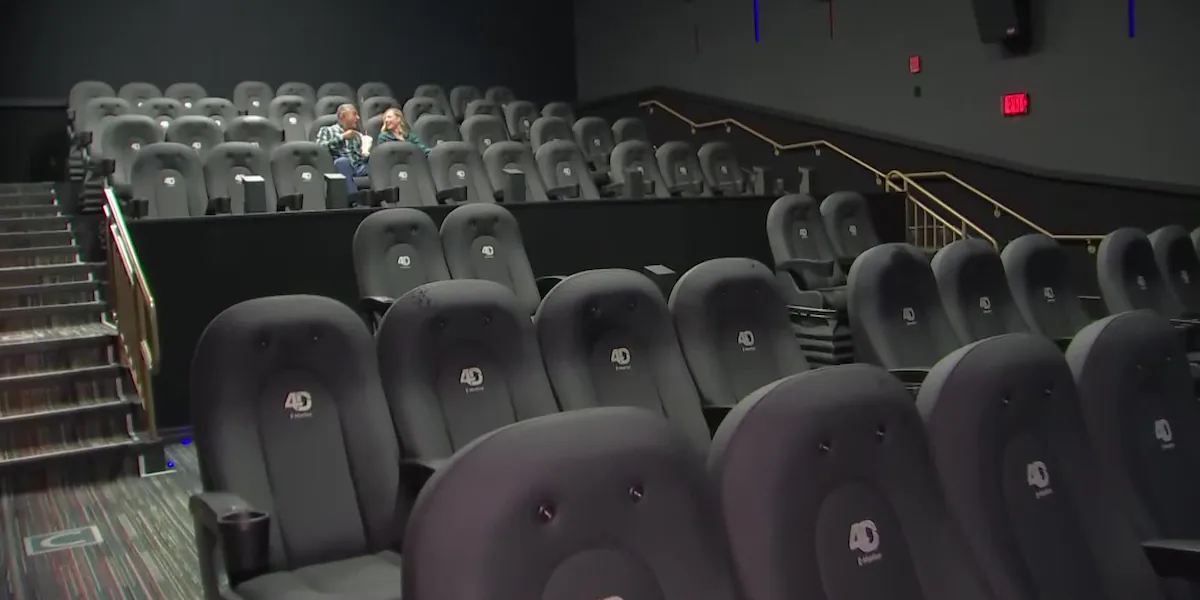 A nearly empty movie theater interior with rows of dark gray motion seats branded with 4D logos and two people seated in the upper tier.