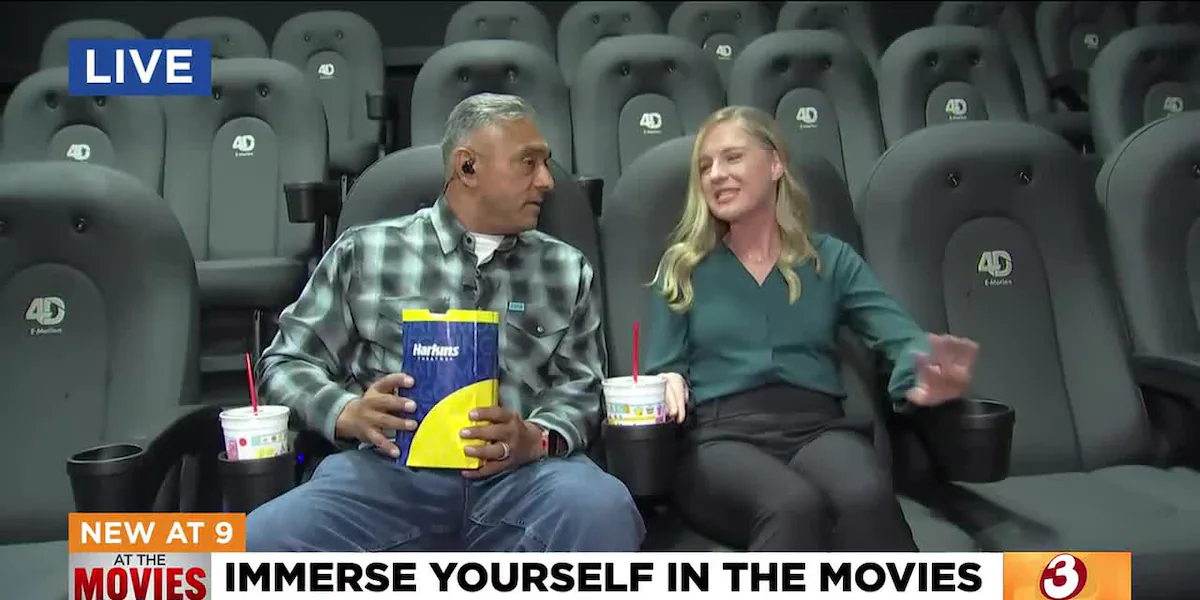Two people sit in branded 4D E-Motion seats inside a Harkins Theatres auditorium holding popcorn and drinks during a live news broadcast.