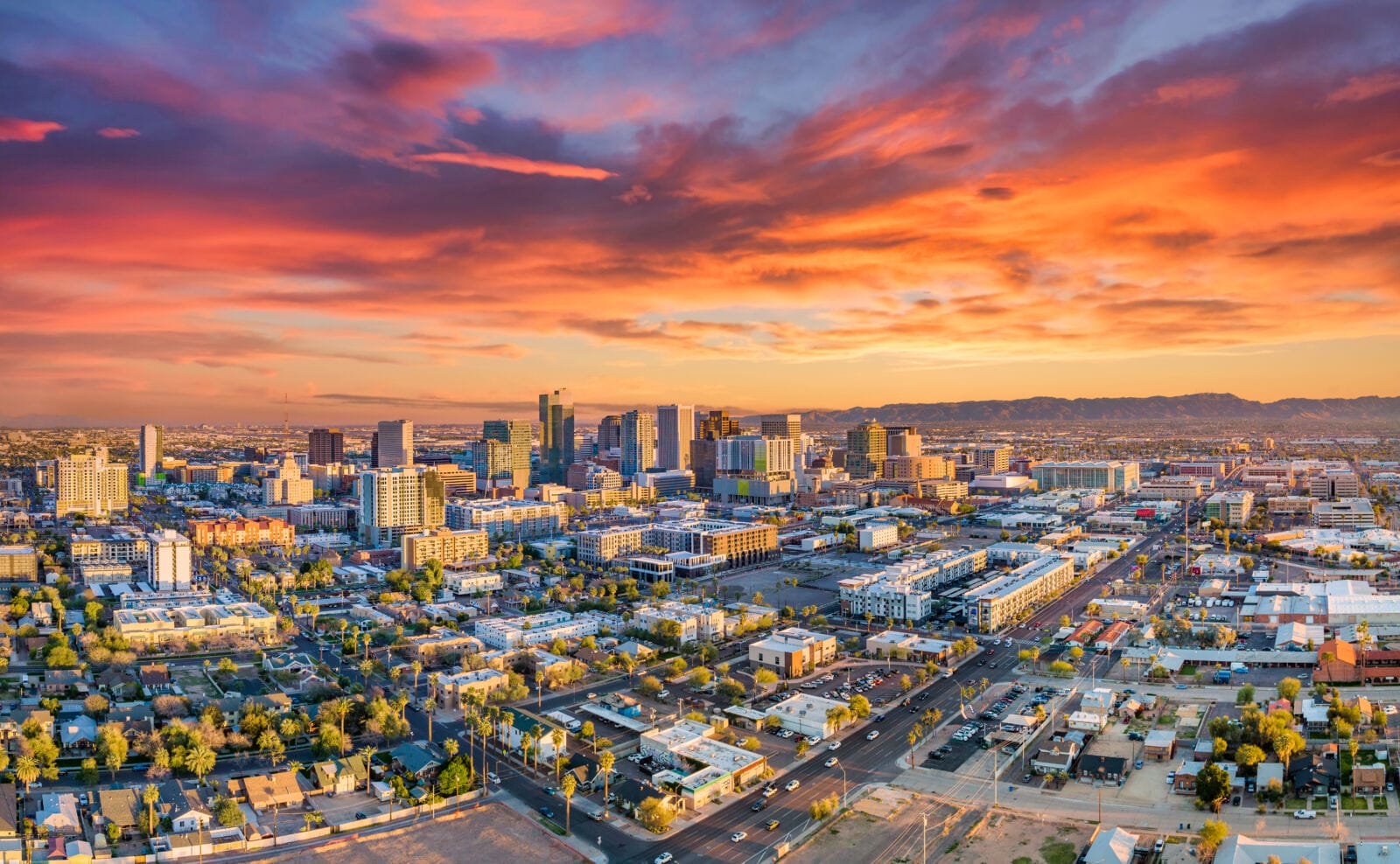 Downtown Phoenix skyline at sunset with palm-tree-lined streets bathed in orange and magenta light, mountains visible in the background