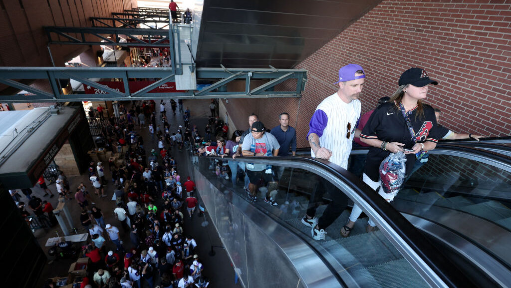 A large crowd of baseball fans fills a stadium concourse at Chase Field, with others riding an escalator upward along a brick wall, several wearing Diamondbacks caps and jerseys.