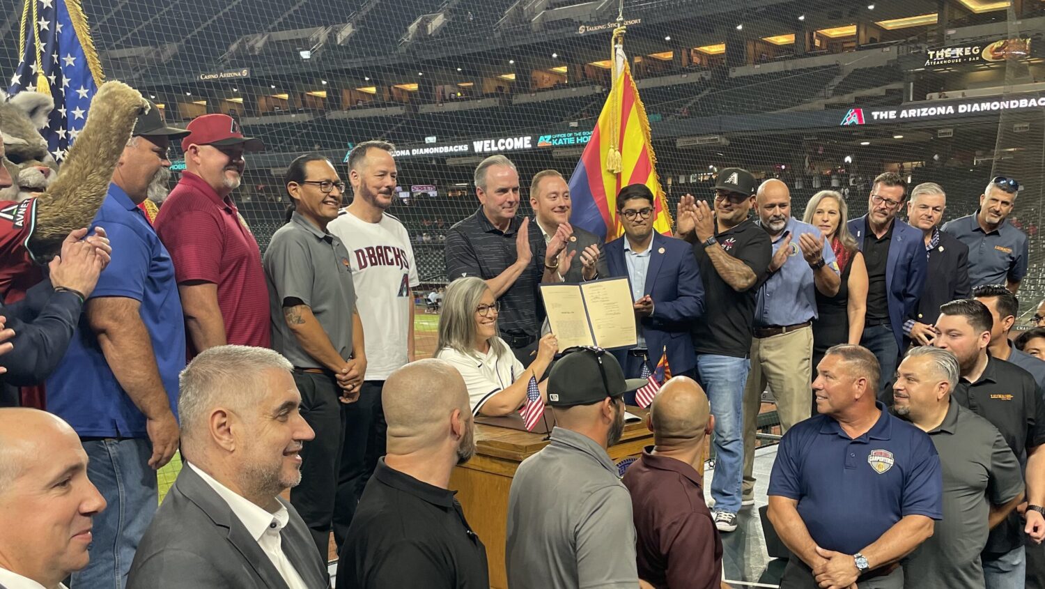 Gov. Katie Hobbs signs HB 2704 at a podium on the Chase Field playing surface, surrounded by applauding officials and the Arizona state flag.