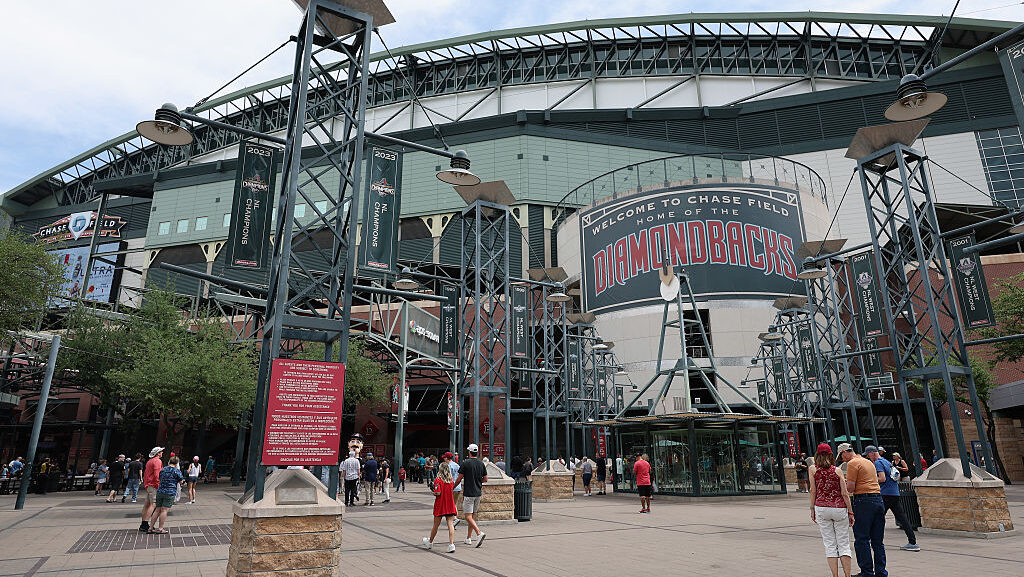 Exterior entrance plaza of Chase Field showing the green steel-frame facade, a Welcome to Chase Field Home of the Diamondbacks sign, 2023 NL Champions banners, and fans walking toward the gates.