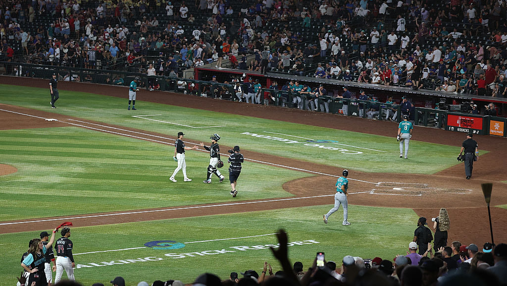 A nighttime MLB game at Chase Field showing players near home plate, Diamondbacks and Mariners uniforms, fans in the stands, and Talking Stick Resort branding on the field.