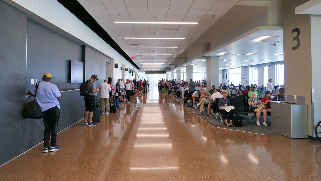 Airport terminal concourse with terrazzo floor, gate 3 sign visible, seated passengers on the right, and travelers walking along the left wall.