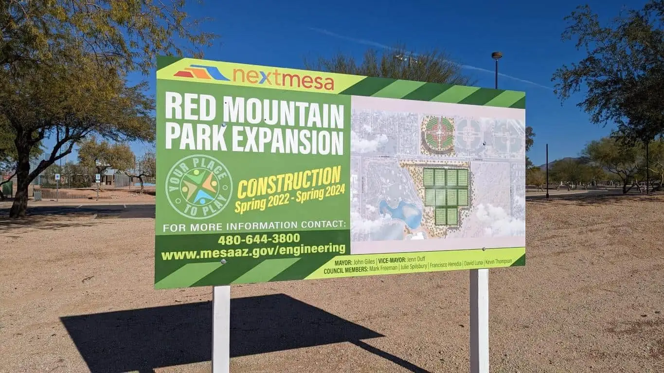 Green construction project sign reading Red Mountain Park Expansion with Spring 2022 to Spring 2024 construction dates, a site plan inset, contact number, and website, standing on white posts in a desert park setting with trees and blue sky.