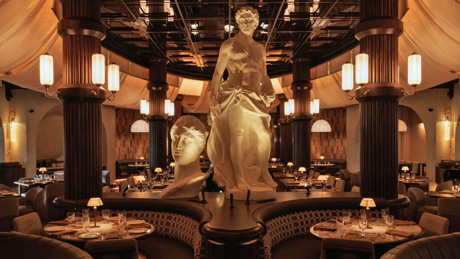 Opulent restaurant interior with two large classical white marble-style sculptures on a raised platform, dark fluted columns, warm pendant and sconce lighting, draped fabric ceiling accents, and formally set dining tables at Roman God of Fire in Tempe