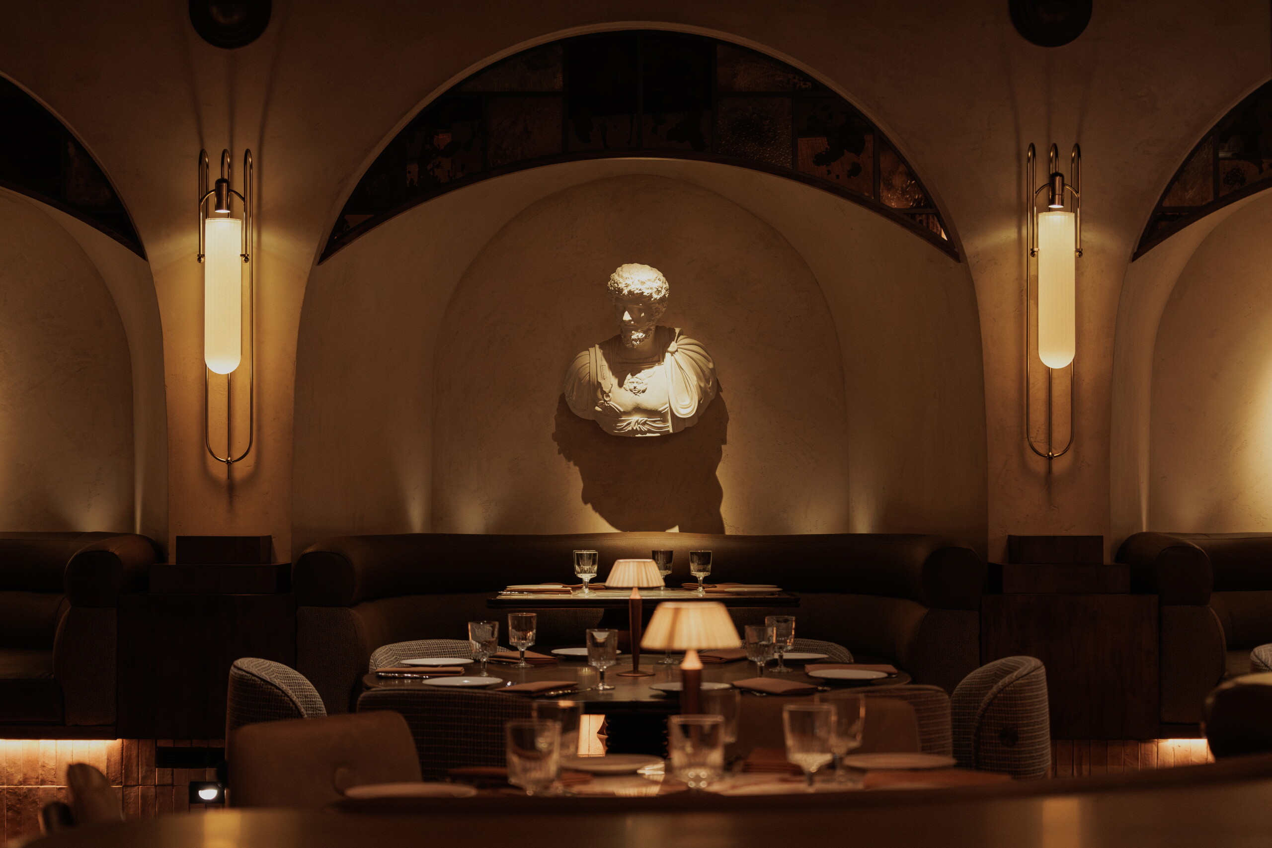 Dimly lit upscale restaurant interior with vaulted arched ceilings, amber wall sconces, dark banquette seating, set tables with glassware, and an illuminated classical Roman-style marble bust in a central wall niche