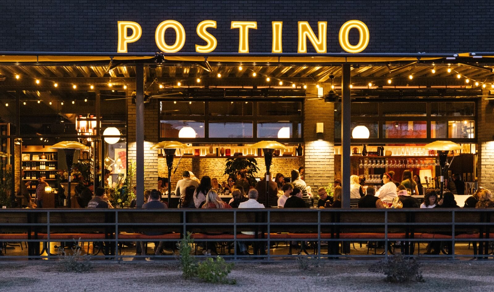 A busy Postino wine bar at night, with a warmly lit open-air patio full of diners under string lights and a large illuminated yellow neon POSTINO sign on a dark brick facade.