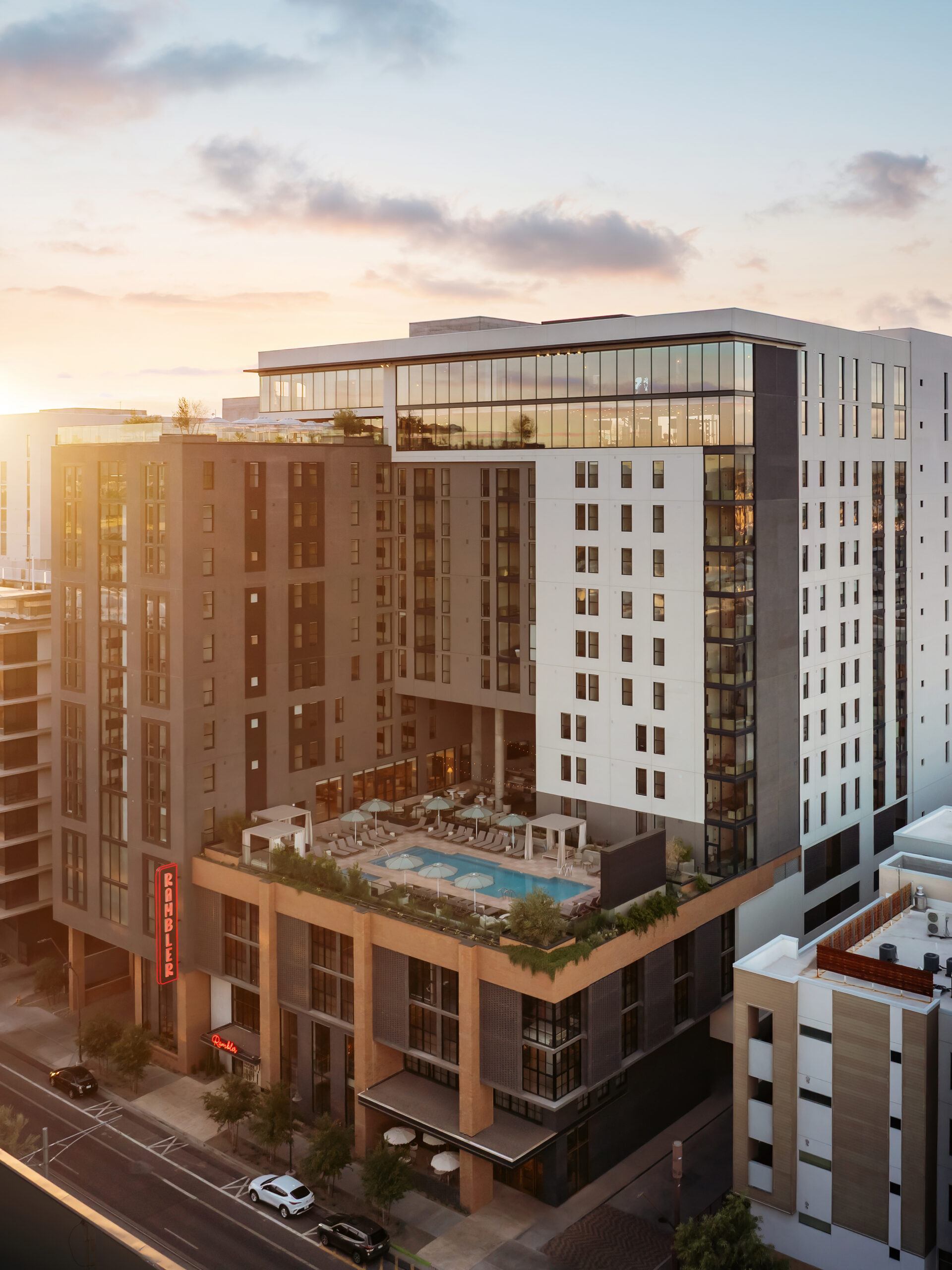 Aerial rendering of the Rambler tower showing a mid-level rooftop pool deck with lounge seating, umbrellas, and Rambler neon signage on the lower facade at golden hour.