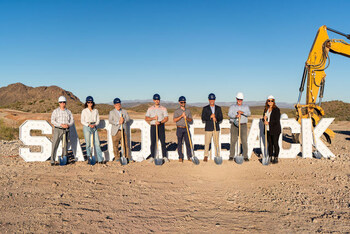 Group of approximately eight people in hard hats holding shovels posing in front of large white SADDLEBACK letters on a desert construction site with a yellow excavator and mountain backdrop visible