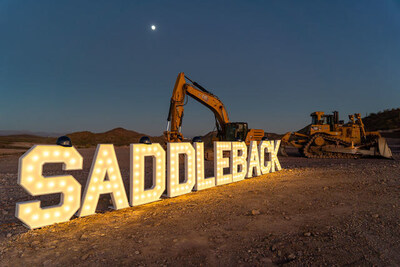 Large illuminated SADDLEBACK marquee sign on a dirt construction site at dusk with a yellow excavator and bulldozer behind it under a twilight sky with a crescent moon