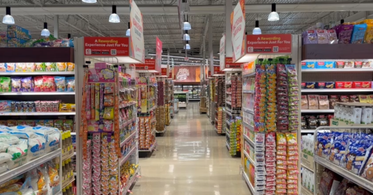 Brightly lit grocery store aisle stocked floor-to-ceiling with Asian snack foods and packaged goods, with red overhead signs reading A Rewarding Experience Just For You visible on both sides of the aisle