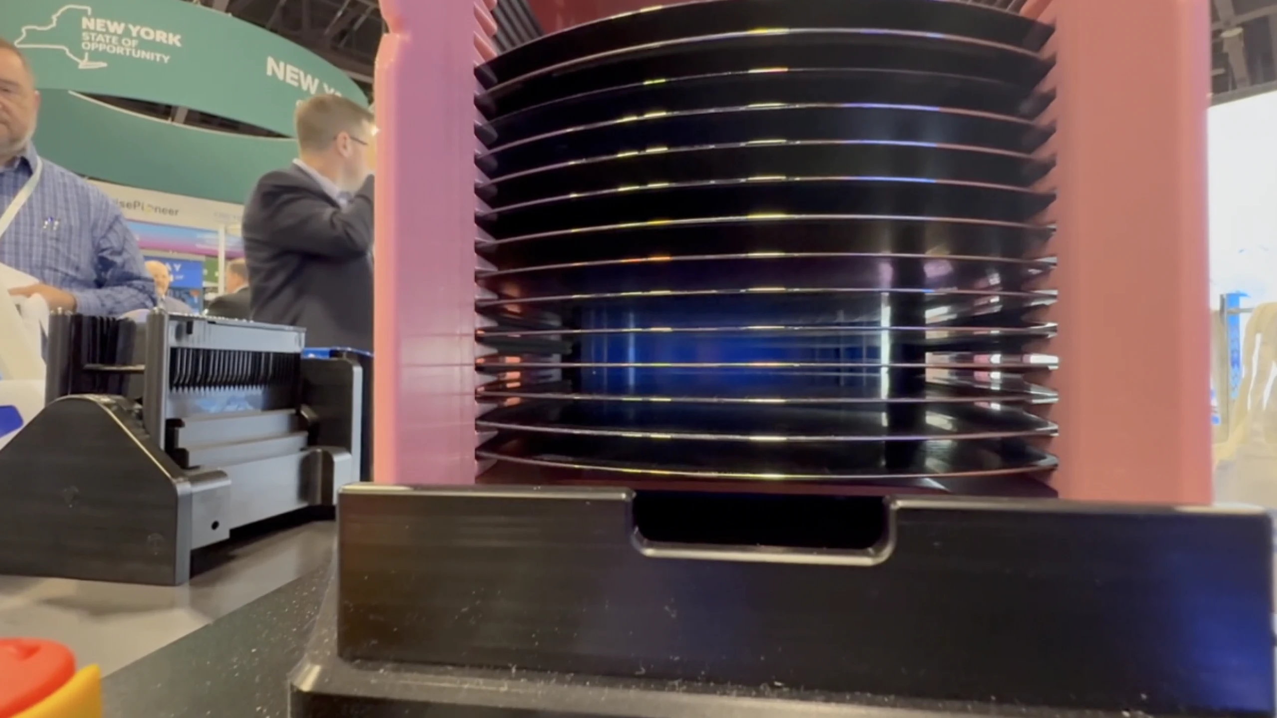 A tall stack of dark semiconductor wafers mounted between pink support columns at a trade show booth, with a New York State of Opportunity banner and attendees visible in the background.