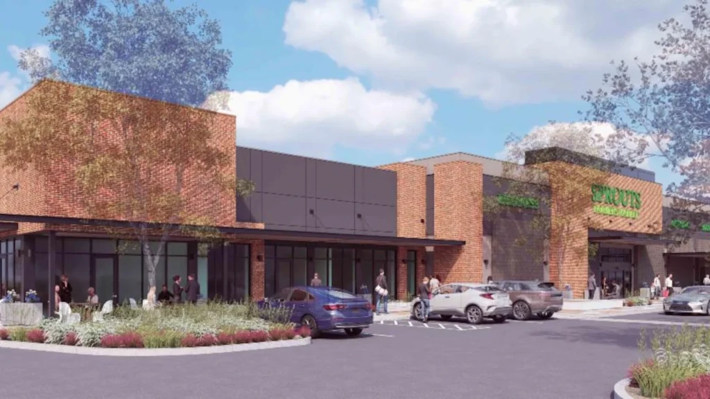 Architectural rendering of Eastmarket at Eastmark showing a strip retail center with brick and dark panel facades, a Sprouts Farmers Market sign on the right anchor unit, shoppers, and parked cars in the foreground.