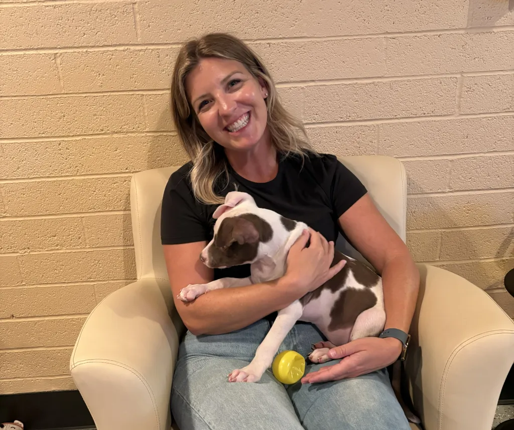 A smiling woman with blonde hair wearing a black t-shirt and jeans sits in a cream armchair holding a brown-and-white puppy, with a yellow ball resting on her lap, photographed against a painted cinder-block wall.