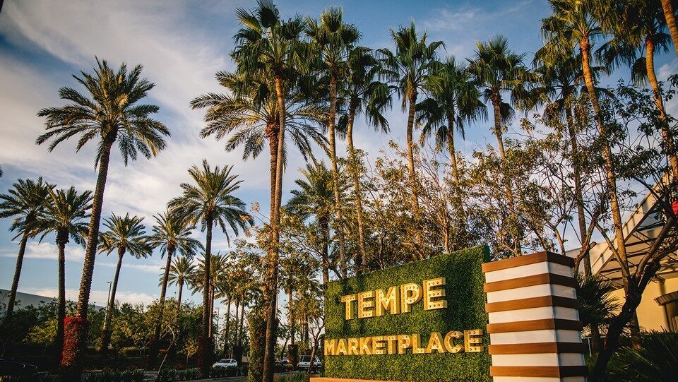 Tempe Marketplace entrance sign with gold letters on a green hedge wall, framed by tall palm trees under a partly cloudy sky.