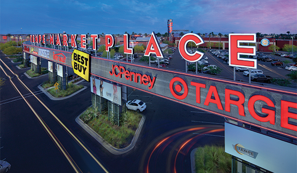 Aerial dusk rendering of Tempe Marketplace showing illuminated rooftop signage spelling TEMPE MARKETPLACE alongside Best Buy, JCPenney, and Target signs over a multi-lane entry road with light trails.