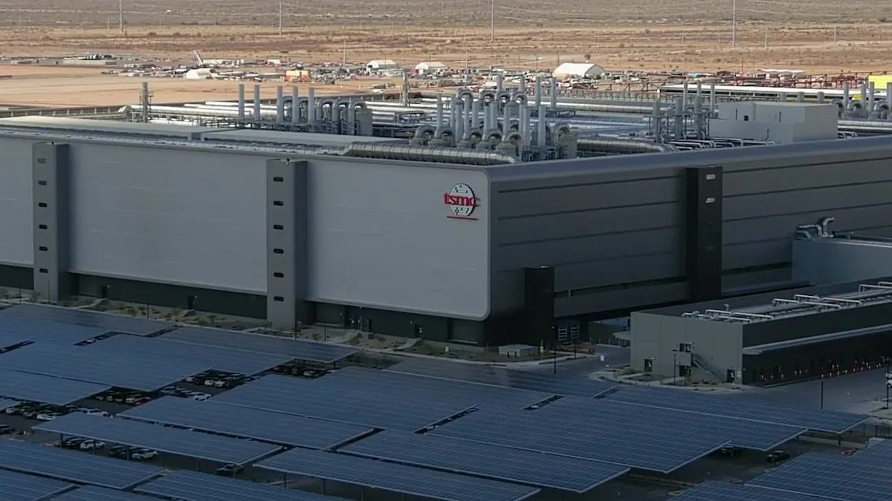 Aerial photo of a completed TSMC fab with the TSMC logo on a gray exterior wall, solar canopy over the parking lot, and pipe infrastructure on the roof