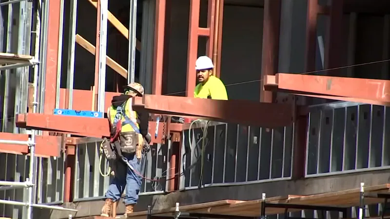 Two construction workers in hard hats and safety harnesses on a multi-story steel-frame building under construction, with red steel beams visible