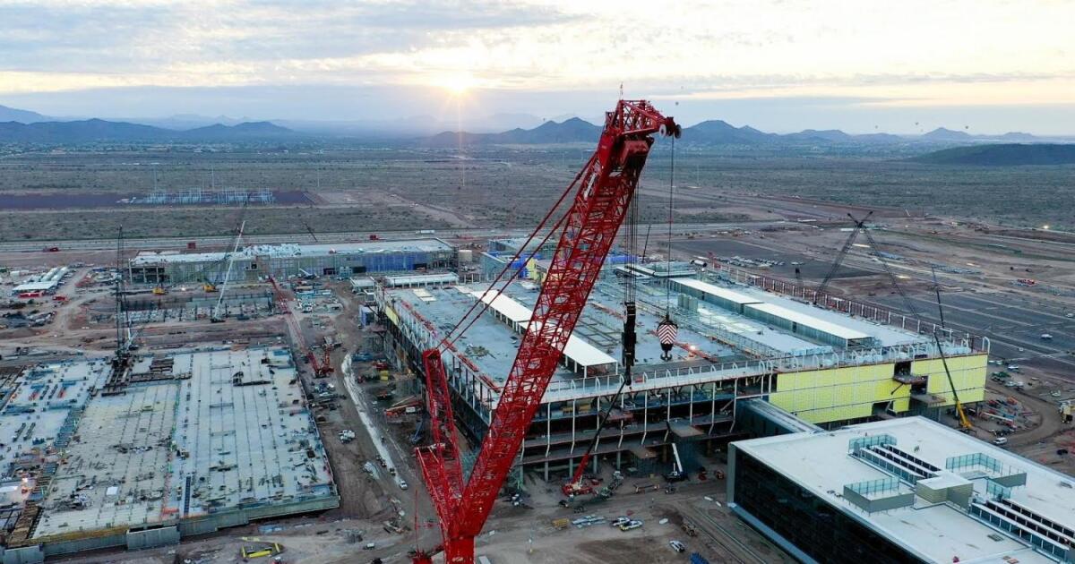 Aerial drone photo of a semiconductor fab under construction in the Arizona desert, with a red crawler crane and yellow-clad steel structures at dusk