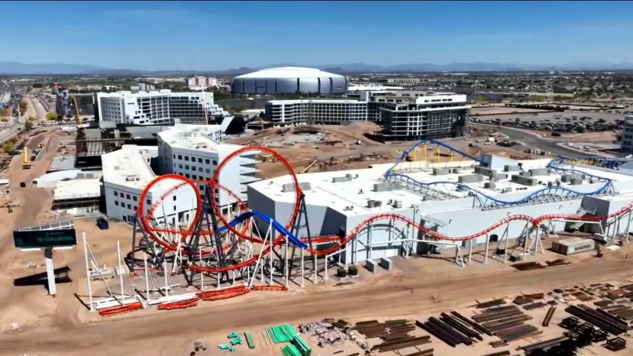 Aerial drone photo of Mattel Adventure Park under construction in Glendale, with red and blue roller coaster loops visible atop white buildings and State Farm Stadium in the background