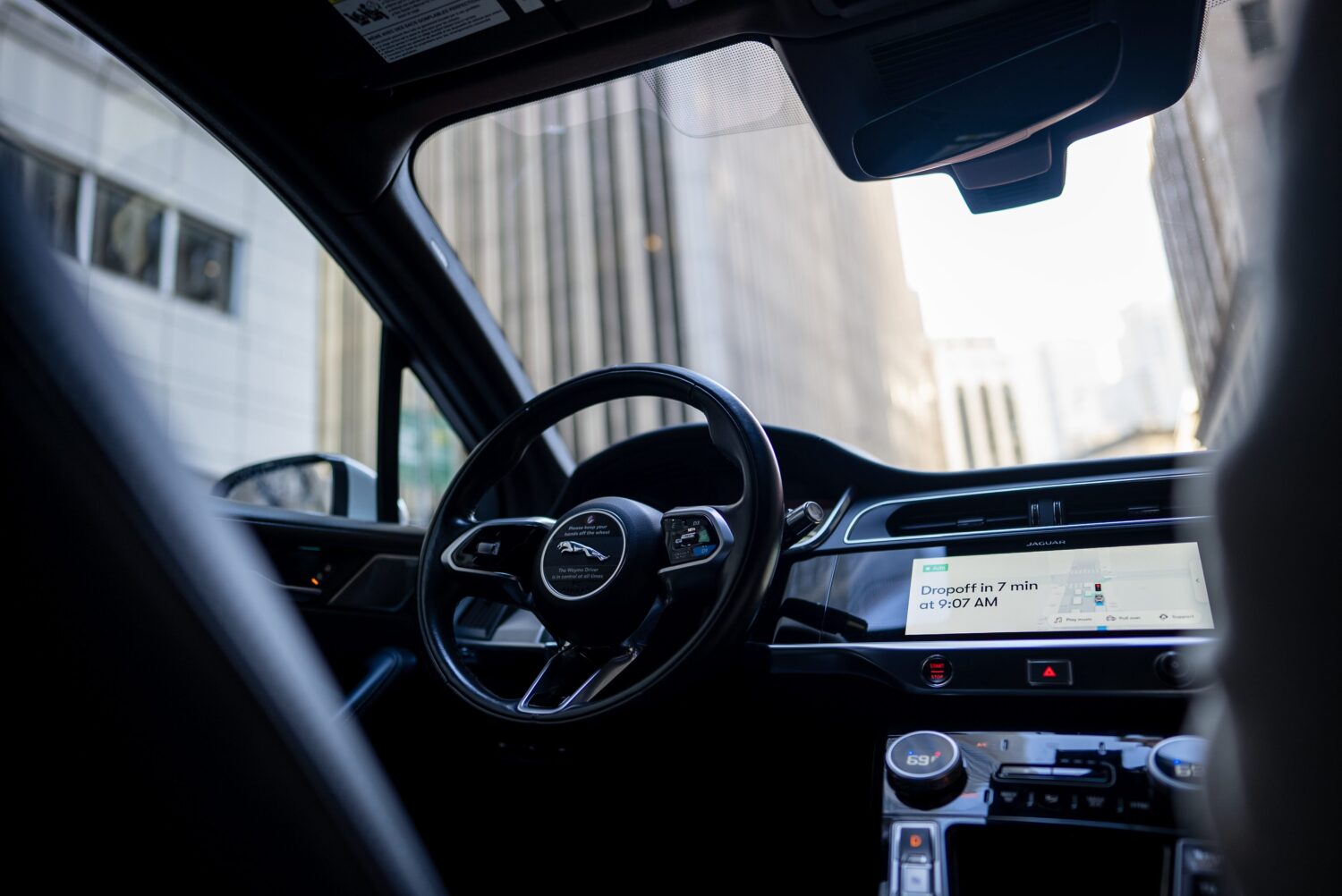 Interior of a Waymo autonomous Jaguar I-PACE robotaxi showing the Waymo-branded steering wheel and an infotainment screen displaying dropoff navigation information