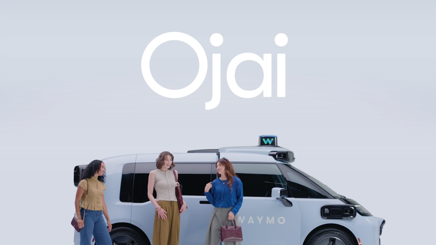 Promotional rendering of three women standing beside a light-blue Waymo Ojai autonomous minivan with a sensor pod on the roof