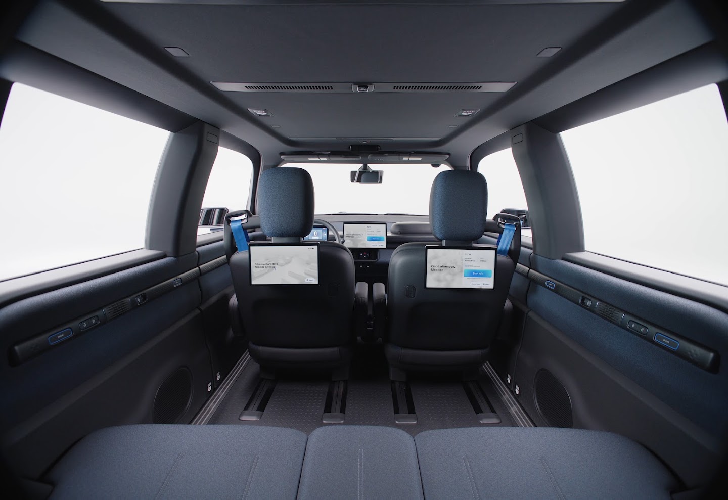 Interior view from rear seat of the Ojai showing two passenger-facing touchscreens mounted on front headrests, blue seatbelts, dark upholstery, and a camera at the top center of the windshield