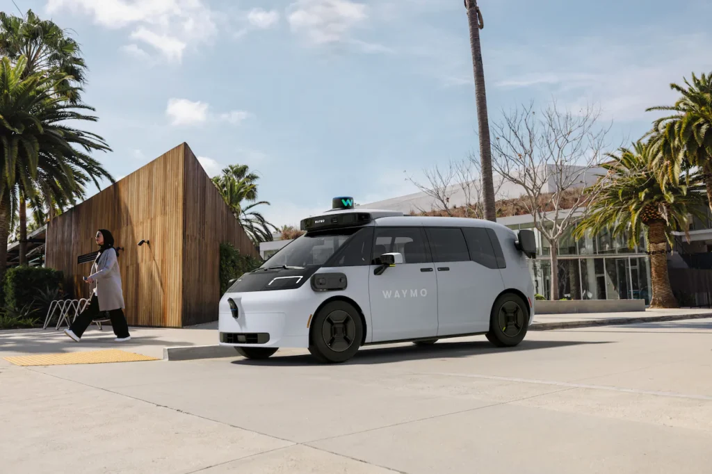 A white Waymo-branded autonomous minivan with a roof-mounted sensor array is parked at a curb in a sunny urban setting with palm trees while a pedestrian walks nearby.